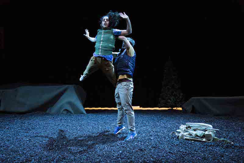 A woman in a green outdoor vest and cargo pants is in mid air after a male dancer throws her. A tent is in the background. Peet and dirt cover the stage floor.